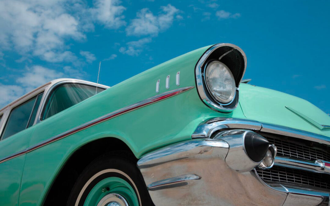 Voiture classique vintage de couleur vert menthe, vue de trois quarts avant. Elle possède des phares ronds et des pare-chocs chromés brillants. Le ciel est bleu avec quelques nuages épars en arrière-plan.